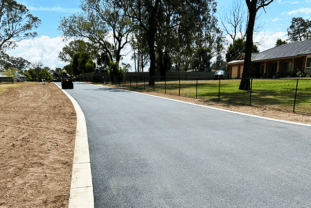 Newly paved asphalt road with trees and residential houses in the background.
